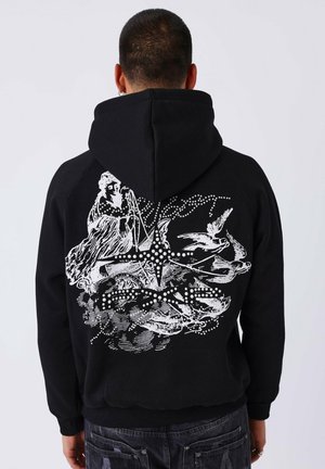 Persona che indossa un hoodie nero con una grafica bianca di una figura alata, una stella e uccelli in volo sulla schiena, in piedi contro uno sfondo semplice.