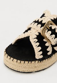 Sandalo nero in suede con dettagli in crema intrecciati, accentuato da una fibbia e una suola piatta in iuta intrecciata. Presenta un design a fascette incrociate.