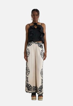 Mujer con blusa negra sin mangas con volantes, pantalones beige de pierna ancha con estampado geométrico negro y sandalias de plataforma, posando contra un fondo liso.