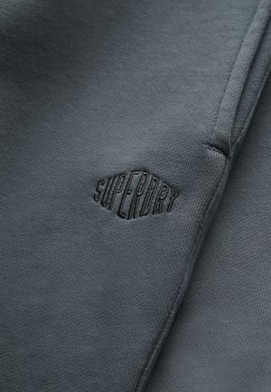 Primer plano de tela gris oscuro con el logo "Superdry" bordado en negro y costura visible.