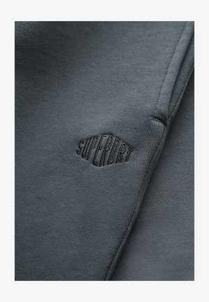 Zbliżenie na ciemnoszary materiał z czarnym wyszywanym logo „Superdry” oraz widocznymi szwami.