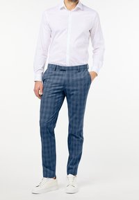 Man med vit långärmad skjorta, blå rutiga byxor, vita sneakers, står med ena handen i fickan mot en enkel vit bakgrund.