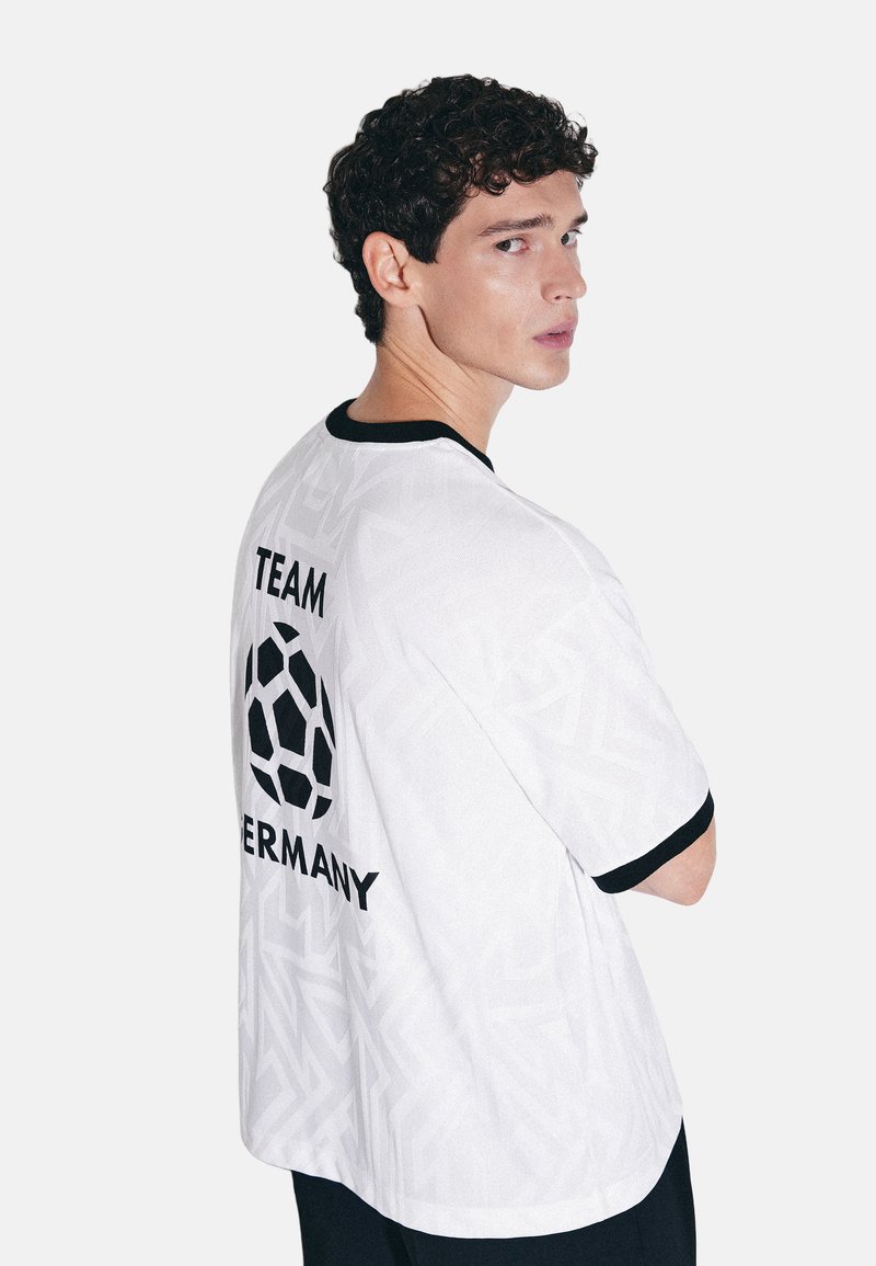 Jeune homme aux cheveux courts et bouclés portant un t-shirt blanc « Team Germany » avec un motif ballon de football noir dans le dos.