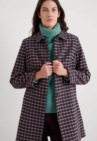 Karo kaput u burgundcu i zelenoj boji, s kopčanjem na dugmad, raširenim kragnom i dugim rukavima, ispod kojeg se nalazi teksturasta zelena majica s visokim ovratnikom.