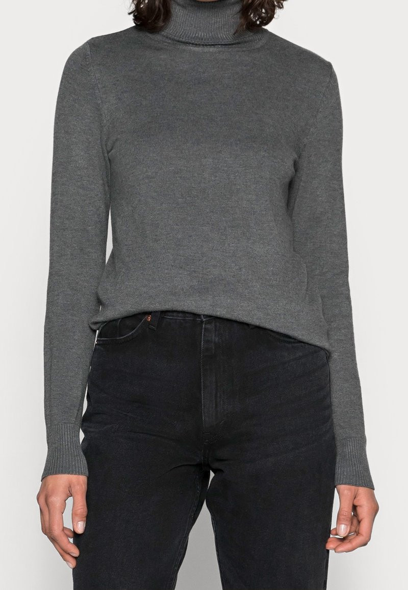 Grå rullekravepullover lavet af blødt strikket materiale med ribstrikkede ærmegab, kombineret med sorte højtaljede jeans, der fremhæver en figursyet silhuet.