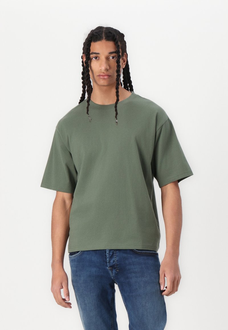 Camisa de gran tamaño en verde oliva con mangas cortas, cuello redondo, hecha de un suave material de algodón, combinada con jeans ajustados de color azul.