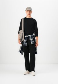 Jeune homme portant une casquette grise, un t-shirt à manches longues noir, un pantalon noir, des chaussures blanches à enfiler, avec une chemise à carreaux nouée à la taille et un sac bandoulière gris.