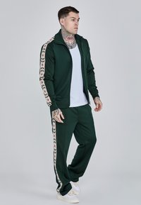 Zelený teplákový súprava s bundou na zips a zodpovedajúcimi nohavicami. Rukávy a bočné strany sú zdobené kvetinovým motívom v béžovej a červenej farbe. Biele tenisky.