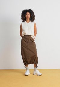 Suéter sin mangas de punto trenzado color crema, falda tipo cargo marrón con lazos ajustables, y zapatillas deportivas blancas. Paleta de colores neutros y terrosos.