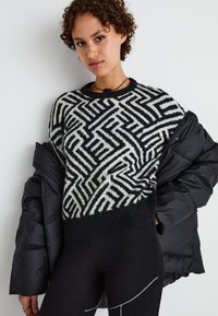Pull en tricot noir et blanc à motif avec un design abstrait en zigzag, col rond et ourlet côtelé, porté avec une veste matelassée noire.