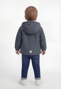 Kleinkind mit kurzen, hellen Haaren, das eine graue Kapuzenjacke mit kleinen Mustern, blaue Jeans und weiße Turnschuhe trägt und mit dem Rücken zur Kamera steht.