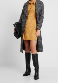 Cappotto lungo a scacchi in bianco e nero su un vestito chemisier in faux suede color senape, abbinato a stivali neri in pelle fino al ginocchio e a una borsa nera strutturata.