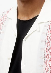 Hvid skjorte med røde broderede mønstre langs knapstolpen. Sort underlag. Knaplukning og blødt, tekstureret stof.