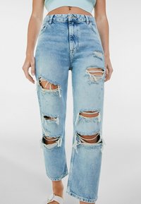 Ljusblå slitna jeans med flera stora hål, fransade kanter och en avslappnad passform. Tyget är denim med en något bleknad finish.