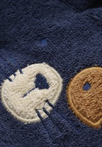 Serviette de bain marine avec une face texturée blanche et une broderie de couleur marron au niveau de l'oreille, ornée de détails en fil contrasté et fabriquée en coton doux.