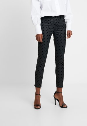 Femme portant un pantalon noir ajusté avec des pois en velours, une chemise blanche à manches longues et des sandales à talons hauts noires sur un fond uni.