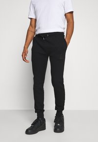 Svarta sweatpants med elastiska manschetter, dragsko i midjan och subtil logodetalj på vänstra låret; kombinerat med en enkel vit t-shirt och svarta sneakers.