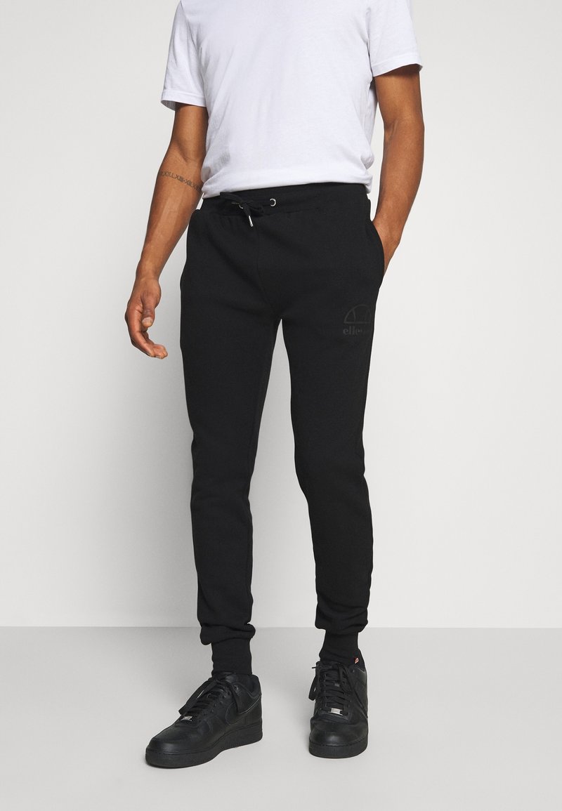 Svarta sweatpants med elastiska manschetter, dragsko i midjan och subtil logodetalj på vänstra låret; kombinerat med en enkel vit t-shirt och svarta sneakers.