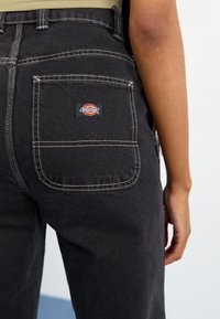 Pantalones de mezclilla negra con costuras blancas, que cuentan con un bolsillo trasero y un parche con el logo de Dickies. Vista en primer plano de la cintura y el área del bolsillo.