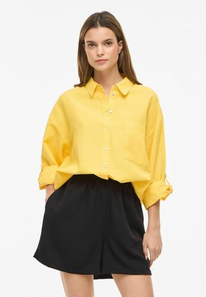 Nő laza, sárga, gombos ingben, felhajtott ujjakkal és fekete, magas derekú rövidnadrágban, egyik kezét a zsebében tartva áll.