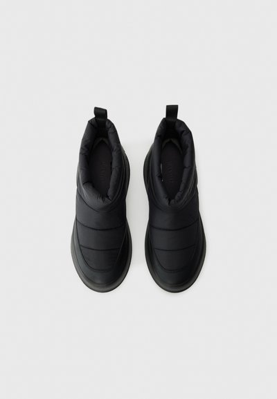 Pár černých izolovaných zimních bot na slip-on s polstrovanými svršky a poutky na zadní straně na jednobarevném bílém pozadí.