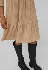 Robe beige côtelée longueur genou avec un ourlet à volants ; associée à des bottes noires montant jusqu'au genou. Présente une coupe décontractée et un tissu texturé.