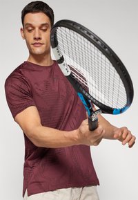 Camiseta deportiva marrón con patrón geométrico, de manga corta y cuello redondo. El modelo sostiene una raqueta de tenis.