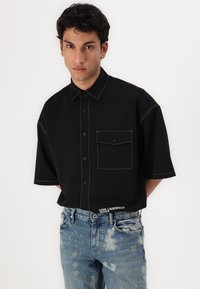 Juodas trumpomis rankovėmis marškinėlis su kontrastuojančiomis baltomis siūlėmis, priekyje yra kišenė ir mygtukų užsegimas; derinama su nusidėvėjusiais mėlynais džinsais, turinčiais pašviesėjusių pleistrų.