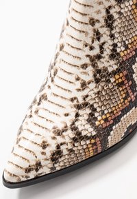 Botín de tobillo de punta afilada hecho de piel de serpiente sintética texturizada, con un patrón de colores blanco, marrón y dorado, y una suela negra elegante.