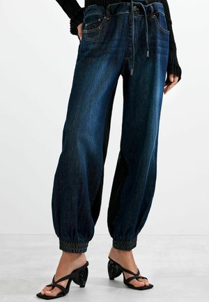 Donkerblauwe denim joggerbroeken met elastische manchetten gedragen met zwarte sandaaltjes met hak en een zwarte top met lange mouwen.
