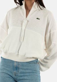 Pólder de forro polar blanco con cremallera de cuarto, puños y dobladillo acanalados, con el logo verde de Lacoste. Textura suave y corte relajado.