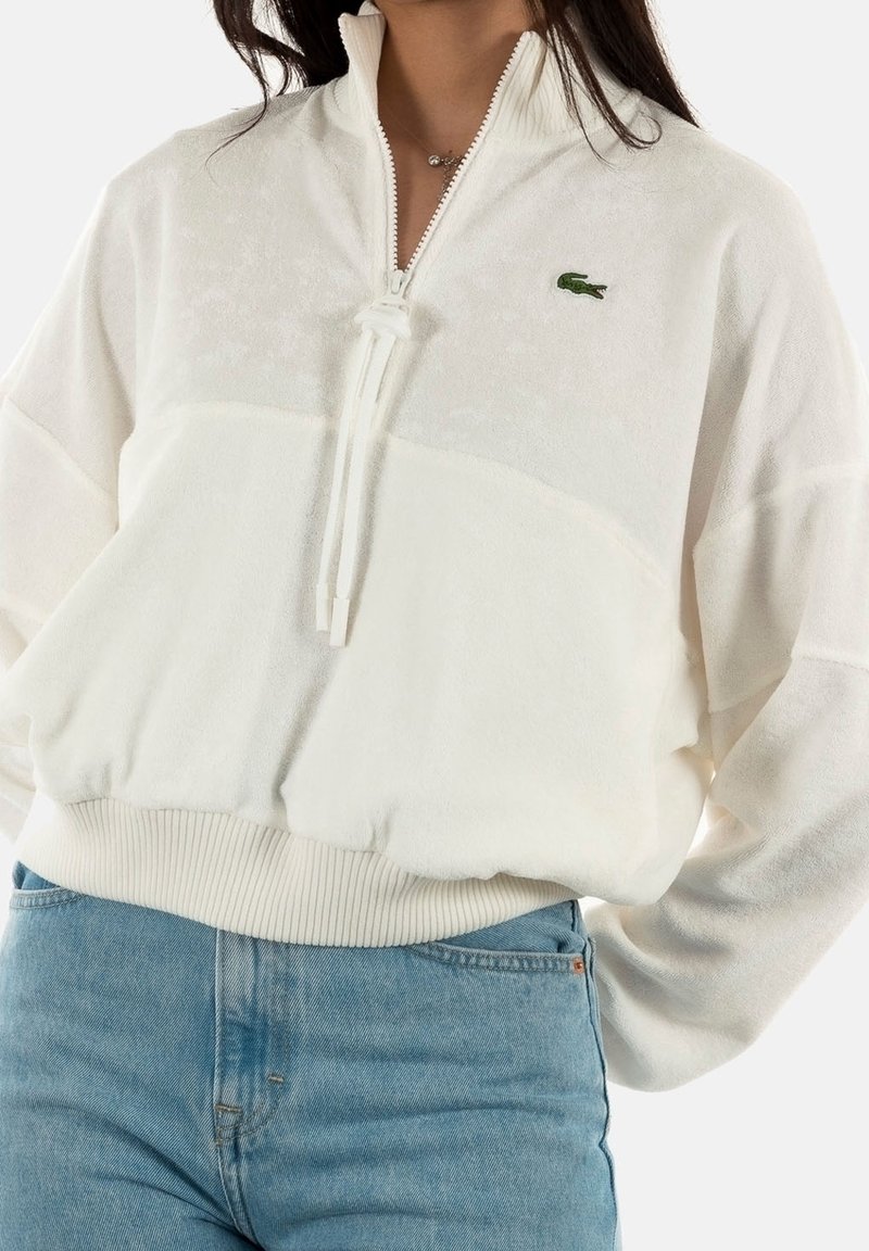 Pólder de forro polar blanco con cremallera de cuarto, puños y dobladillo acanalados, con el logo verde de Lacoste. Textura suave y corte relajado.