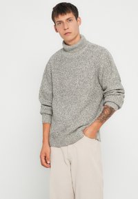 Maglione grigio a collo alto lavorato a maglia con motivo testurizzato, vestibilità ampia e polsini a costine. Indossato sopra pantaloni in corduroy beige.