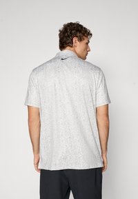 Kortærmet polo t-shirt i lys grå med blomsterdesign, der har krave og et lille sort logo på ryggen.