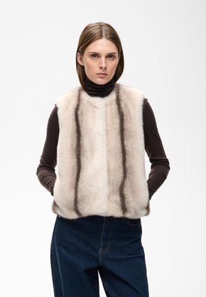 Femme portant un gilet en fausse fourrure beige clair avec des rayures verticales foncées par-dessus un col roulé noir à manches longues et un jean bleu, debout les mains dans les poches.
