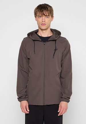 Jonge man draagt een bruin capuchonjack met ritssluiting met zwarte trekkoorden en een zwarte broek, staand tegen een effen lichte achtergrond.