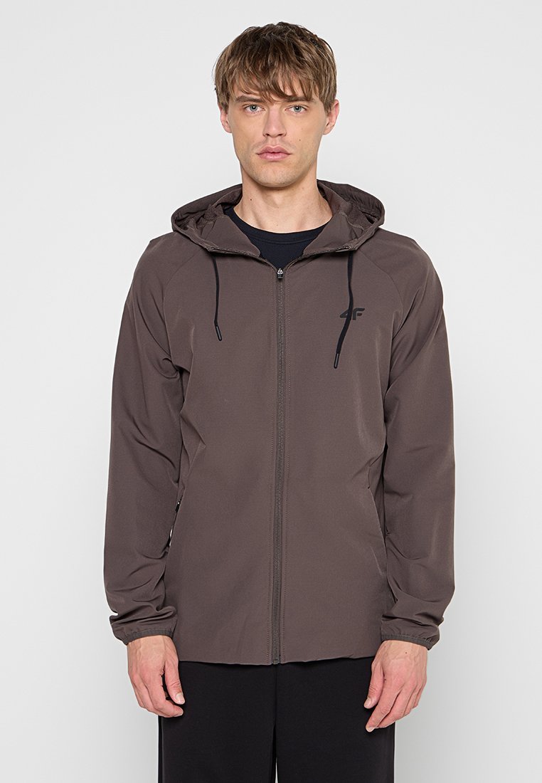 Jeune homme portant une veste à capuche marron zippée avec des cordons noirs et un pantalon noir, debout devant un fond clair uni.