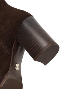 Bruine suède enkellaars met een chunky houten hak. Bevat soepele lijnen, gestikte details en een kleine decoratieve hardware-accent.