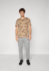 Beige t-skjorte med kamuflasjemønster og korte ermer, med en liten logo, kombinert med lysegrå joggebukser med hvit logo, brukt med svarte joggesko.