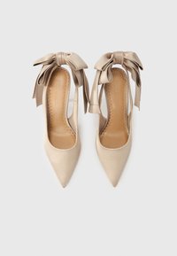 Μπεζ σατέν ψηλοτάκουνα παπούτσια με μυτερή μύτη και λουράκια στον αστράγαλο με μεγάλους φιόγκους, με εσωτερικές σόλες με κυματιστό σχέδιο και ανάγλυφη την επιγραφή "Steve Madden".