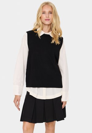 Black sleeveless sweater over a white button-up shirt, paired with a black pleated skirt. Smooth texture, ribbed hem, and classic layering.