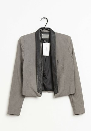 Grauer kurzer Blazer mit schwarzen Satin-Revers, an einem schwarzen Bügel vor weißem Hintergrund hängend.