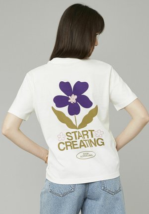 T-shirt en coton blanc avec un grand graphisme de fleur violette entourée de feuilles vertes et le texte "COMMENCEZ À CRÉER" en or, avec des accents rose clair.