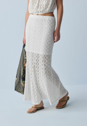 Frau trägt einen weißen Spitzen-Maxirock und ein kurzes Top, hält eine gemusterte Tragetasche, trägt braune Flip-Flops auf hellblauem Hintergrund.