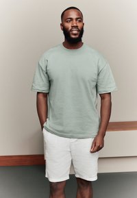 Groen kortärmelig T-shirt van katoen, met een ronde halslijn en een losse pasvorm, gecombineerd met witte shorts tegen een neutrale achtergrond.