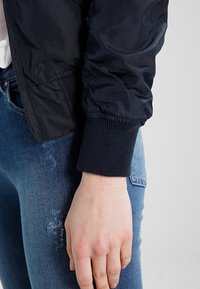 Granatowa kurtka bombowiec o gładkiej powierzchni, z prążkowanym mankietem i elastycznym dołem. Noszona na podartych niebieskich dżinsach. Tekstura jest gładka i lekko błyszcząca.