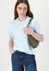 Camiseta de algodón azul claro con mangas cortas, combinada con jeans azul oscuro. El modelo sostiene un bolso de hombro verde oliva con forma curva.