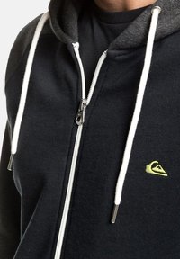 Sudadera con cremallera color marino, con capucha gris y cordones blancos. Presenta una cremallera plateada y un pequeño logo de la marca en amarillo en el pecho.