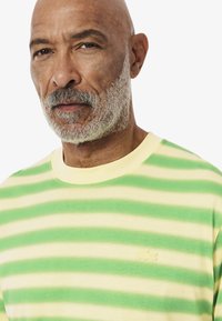 Gestreepte T-shirt met korte mouwen in groen en geel. Heeft een ronde hals en een klein geborduurd logo aan de linkerkant van de borst. Zachte textuur.