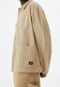 Lysebrun bomuldsjakke med en afslappet pasform, trykknapslukning, en stor forlomme og et sort mærke ved kanten, båret over matchende lysebrune bukser.